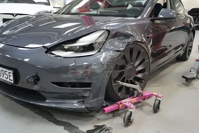 Grauer Tesla Model 3 mit deformiertem vorderem Kotflügel, aufgebockt auf Rangierrollen in einer Werkstatthalle