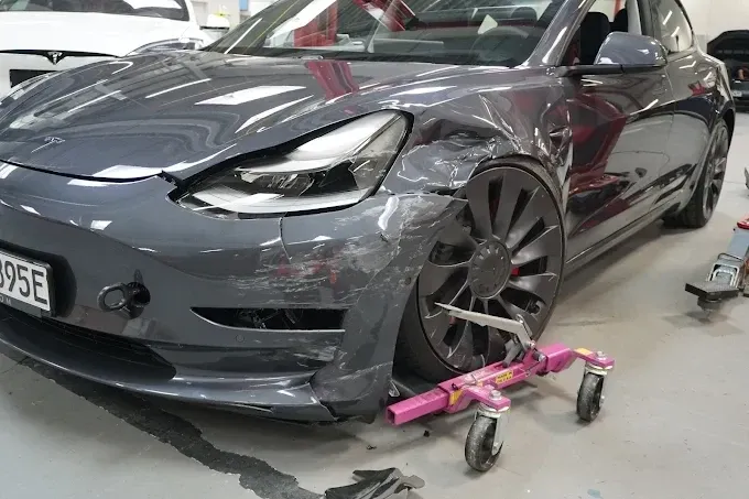 Grauer Tesla Model 3 mit deformiertem vorderem Kotflügel, aufgebockt auf Rangierrollen in einer Werkstatthalle